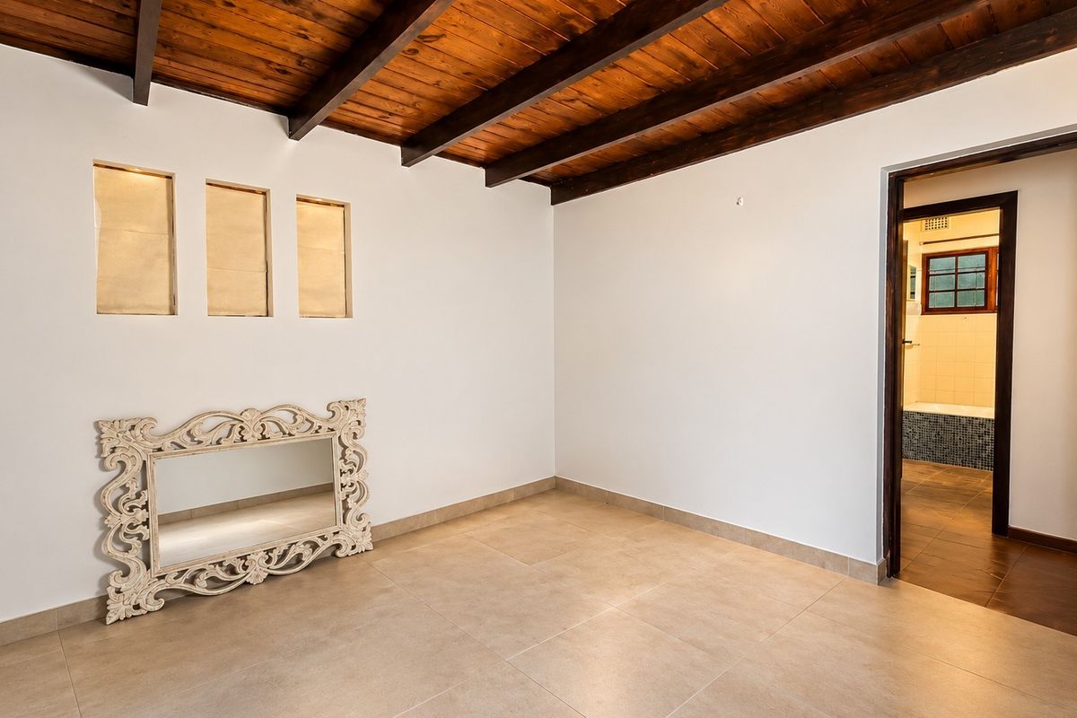 A bright, empty bedroom featuring exposed timber ceiling beams, neutral tiled flooring, and direct access to an adjoining bathroom.