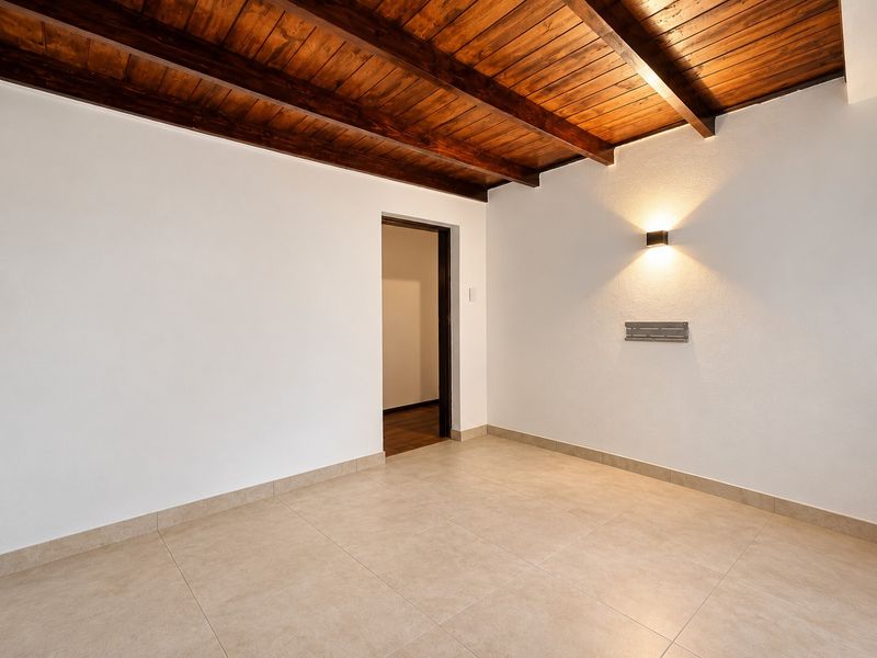 A bright, empty living area featuring attractive exposed timber ceiling beams, neutral tiled flooring, and modern wall-mounted lighting.