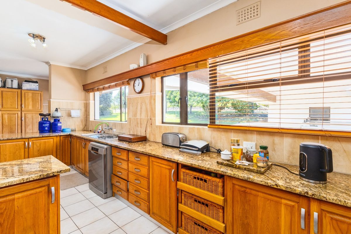 A functional kitchen featuring warm timber cabinetry, granite countertops, tiled flooring, and large windows with venetian blinds overlooking the farm grounds.