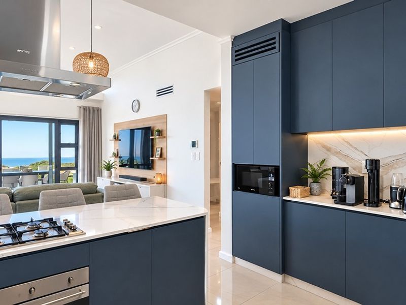 Modern open-plan kitchen and living area featuring dark blue cabinetry, stone countertops, integrated appliances, and seamless flow to a balcony with ocean views.