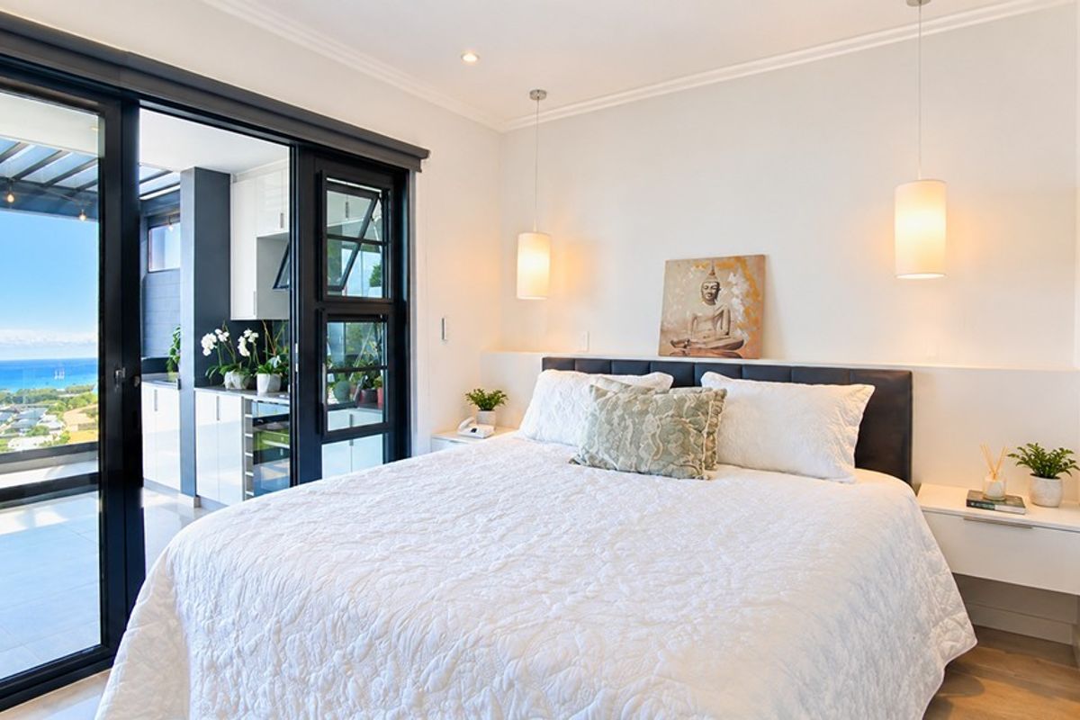 A bright, modern bedroom featuring hardwood floors, elegant pendant lighting, and large sliding glass doors that open onto a balcony with scenic coastal views.