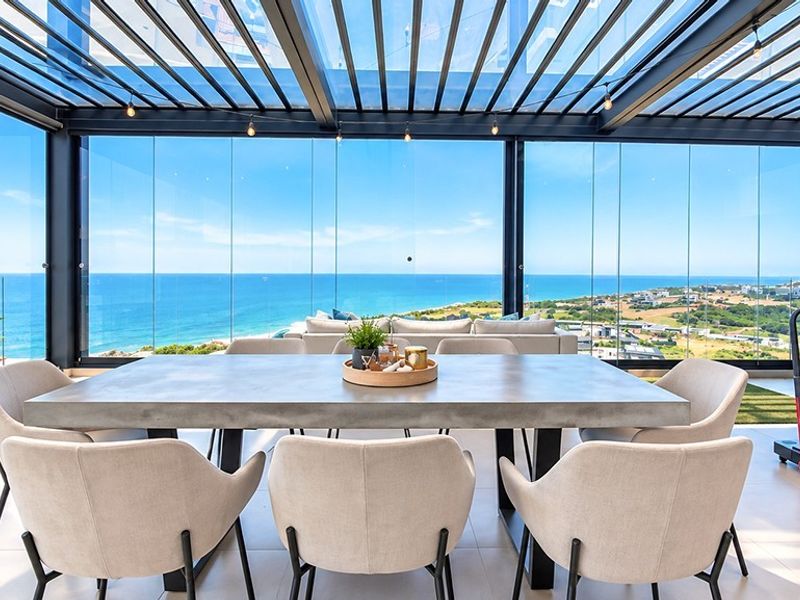 A luxurious enclosed balcony featuring a retractable louvered roof, frameless glass walls, and panoramic ocean views, perfect for year-round outdoor dining and relaxation.