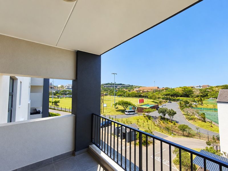 A modern, covered balcony featuring tiled flooring and a sleek metal balustrade, offering expansive views over the surrounding estate and recreational grounds.