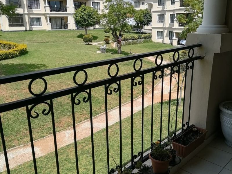 A covered balcony featuring a decorative wrought iron balustrade and tiled flooring, overlooking a well-maintained communal garden area within the apartment complex.