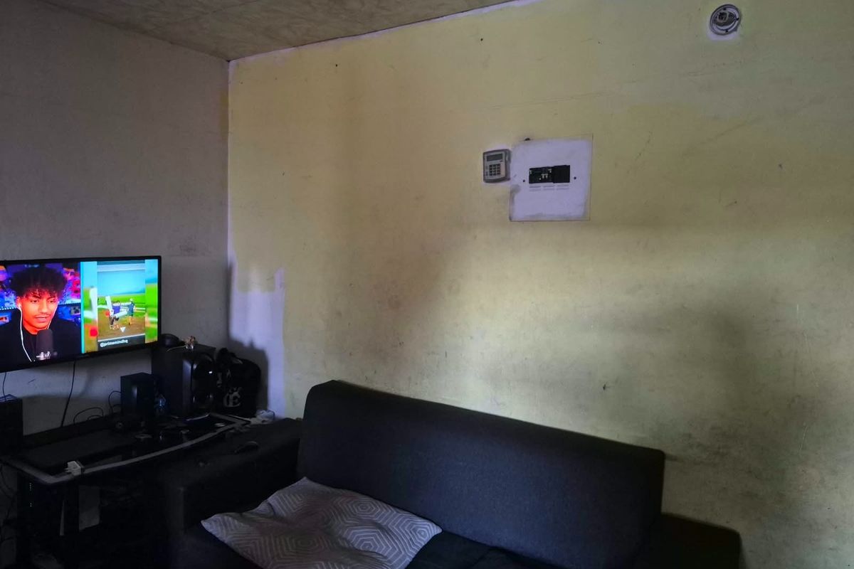 A living room area featuring a wall-mounted security keypad and electrical distribution board, currently requiring cosmetic updates to the wall finishes.