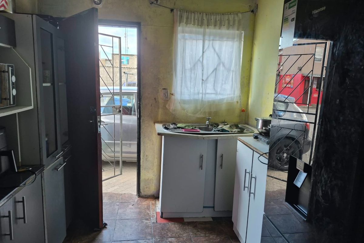 A compact kitchen area featuring basic cabinetry, a sink, and tiled flooring, with direct access to the exterior through a security gate.