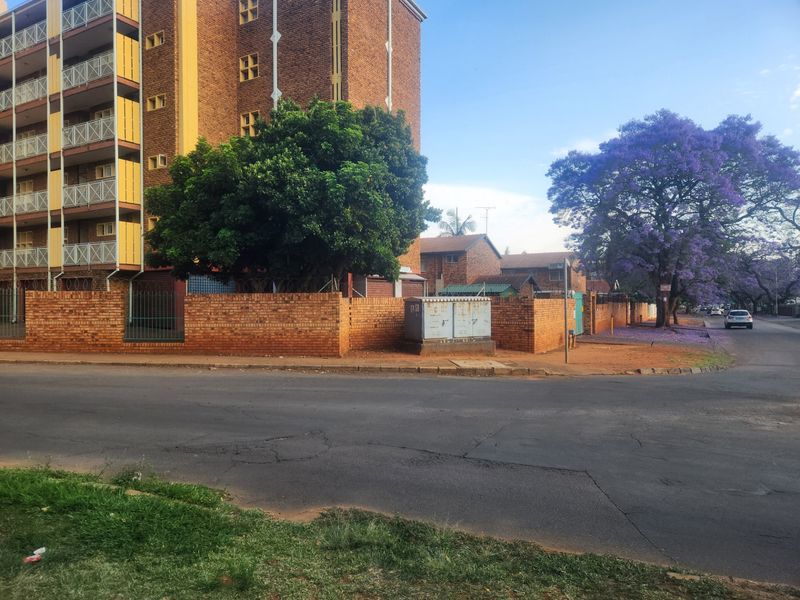 Street view of a multi-story apartment complex featuring brick facade, security fencing, and mature trees along the roadside.