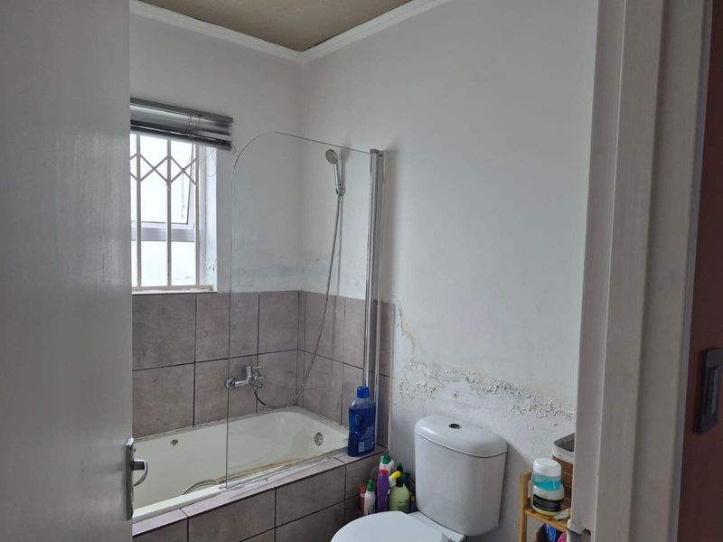 A compact bathroom featuring a bathtub with a shower screen, toilet, and window with security bars. The walls show visible signs of moisture damage.