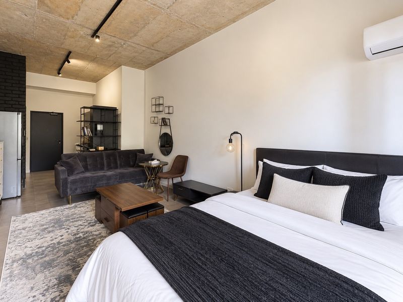 Modern open-plan studio apartment featuring an exposed concrete ceiling, sleek tiled flooring, a black brick accent wall, and a wall-mounted air conditioning unit.