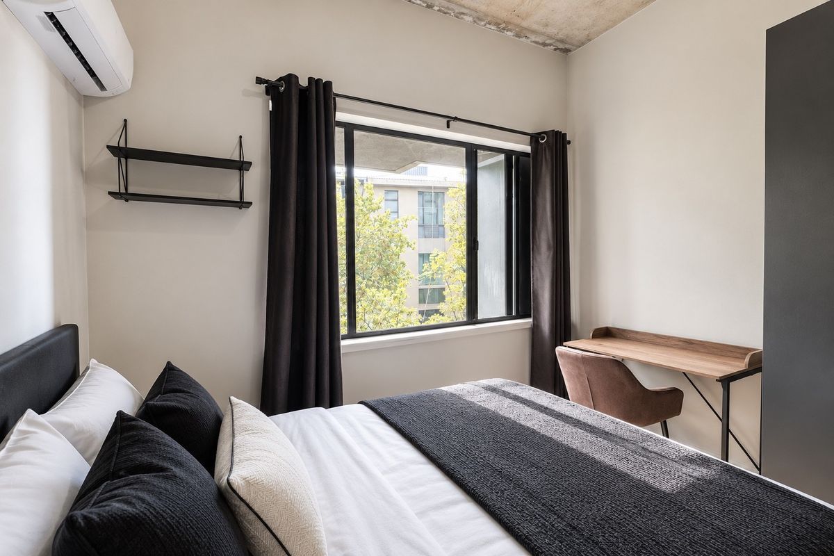 A modern bedroom featuring an exposed concrete ceiling, air conditioning unit, and a built-in wardrobe with a view of the surrounding neighborhood.