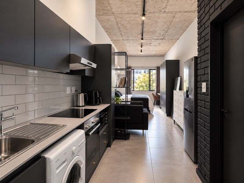 Modern open-plan apartment featuring a sleek kitchen with dark cabinetry, subway tile backsplash, exposed concrete ceilings, and integrated appliances, flowing into a bedroom area.
