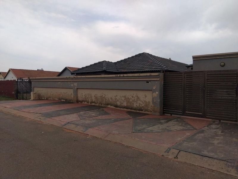 Front exterior view of the property featuring a paved driveway, metal gate, and a perimeter wall requiring maintenance and repainting.