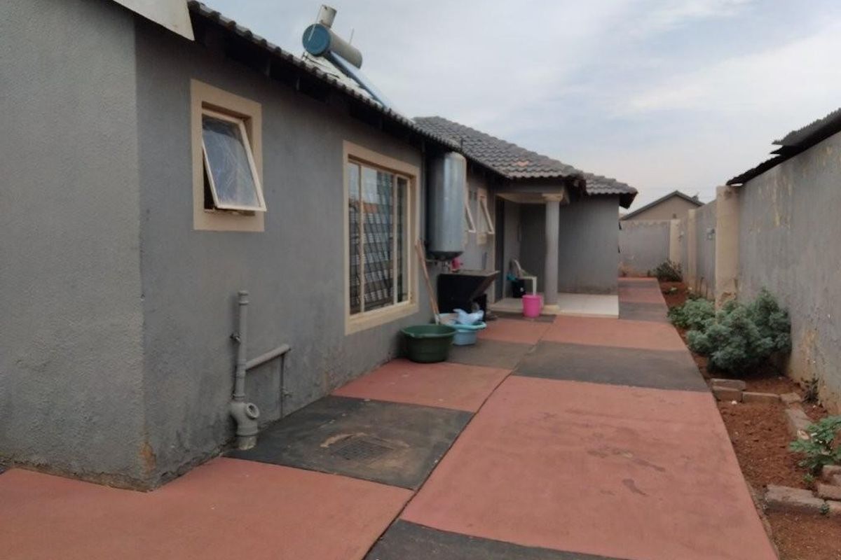 Rear exterior view of the house featuring a paved walkway, solar water heating system, and boundary wall enclosure.