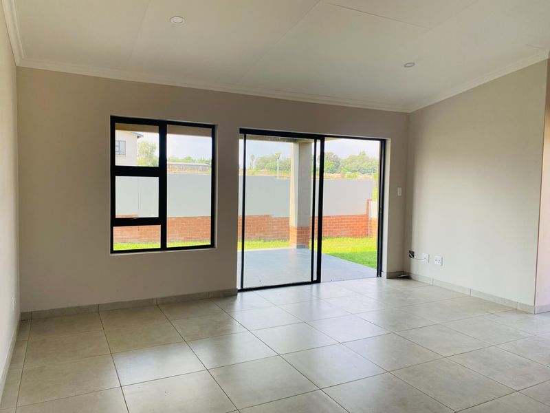 A bright, empty living room featuring neutral tiled floors, modern black aluminium window and sliding door frames, and recessed ceiling lighting.