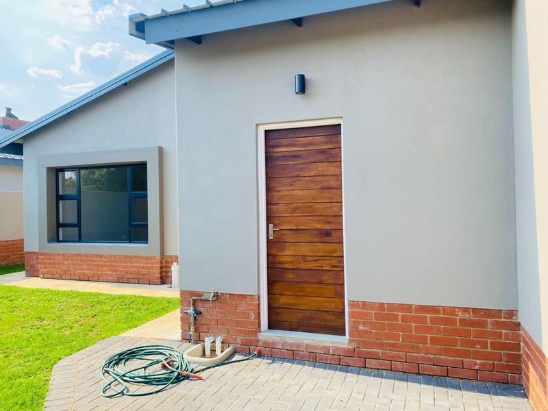 A well-maintained rear exterior view featuring a modern timber door, brick and plaster facade, aluminium window frames, and a paved patio area.