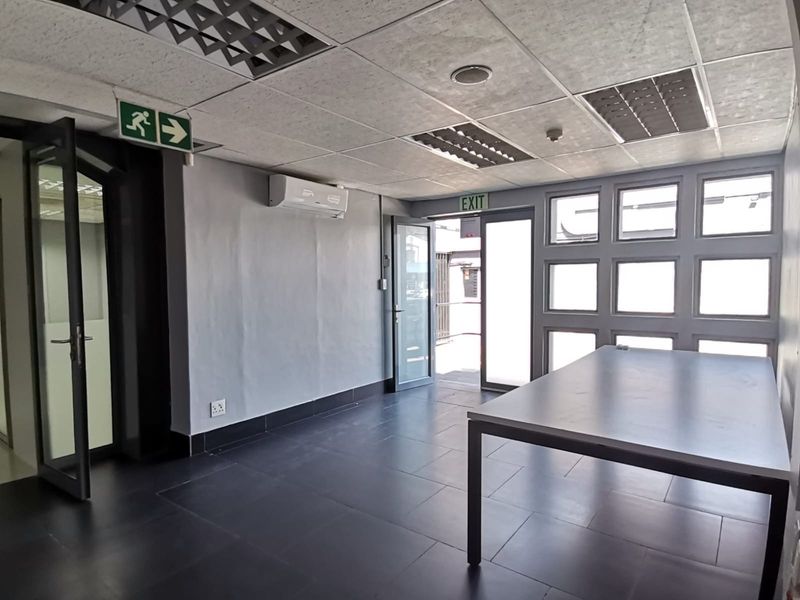 A bright, open-plan office space featuring tiled flooring, a suspended ceiling with recessed lighting, and a wall-mounted air conditioning unit.