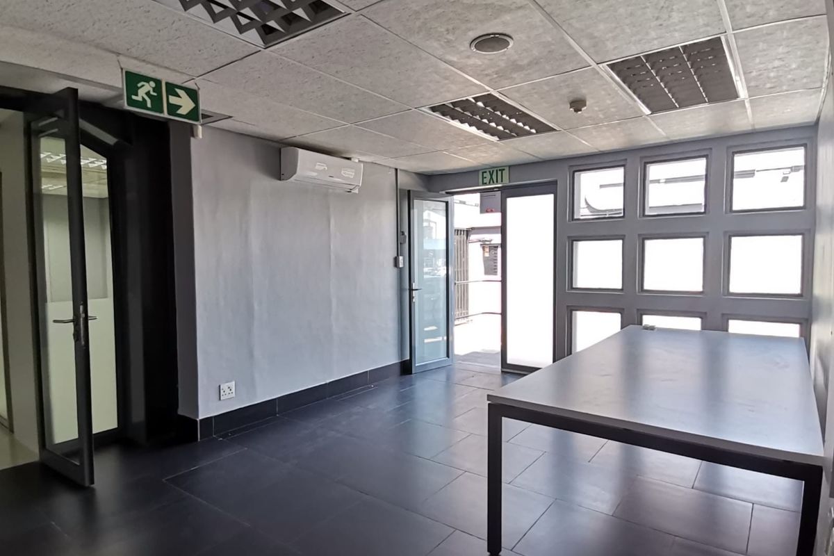 A bright, open-plan office space featuring tiled flooring, a suspended ceiling with recessed lighting, and a wall-mounted air conditioning unit.