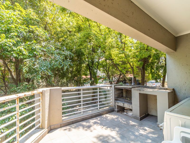 Covered balcony featuring a built-in braai, tiled flooring, and metal balustrade, overlooking a lush garden area.