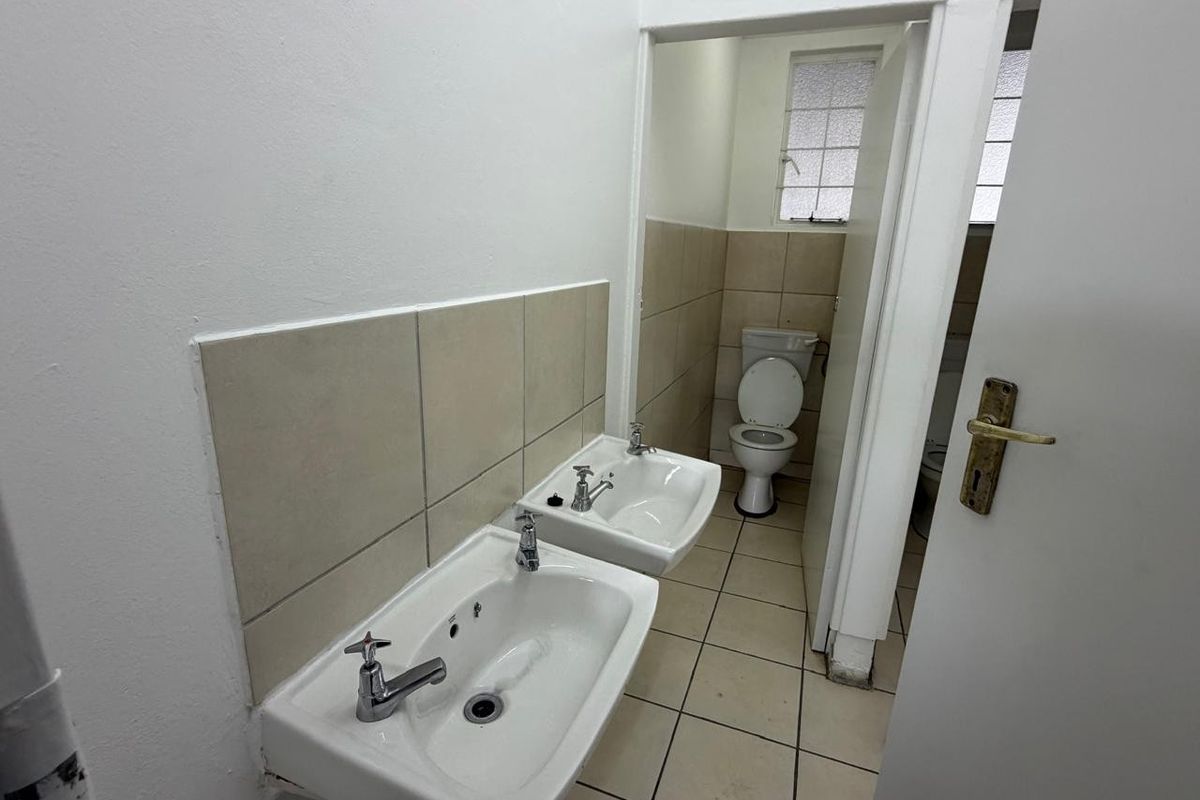 Functional industrial bathroom featuring dual wall-mounted basins, tiled flooring, and a separate toilet cubicle with natural light from an obscured glass window.