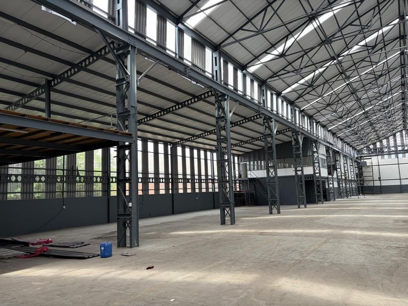 Expansive industrial warehouse space featuring high ceilings, exposed steel structural framework, mezzanine level, and abundant natural light through clerestory windows.