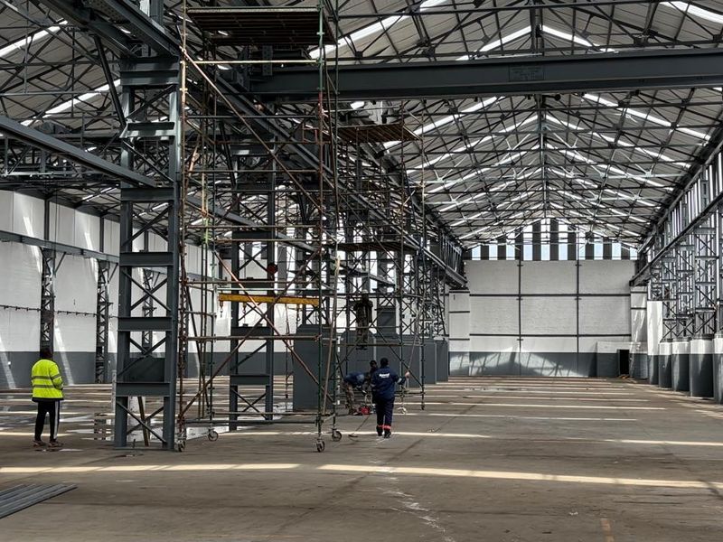 Large industrial warehouse space featuring high ceilings with exposed steel trusses, concrete flooring, and natural light from overhead skylights.