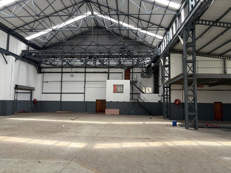 Spacious industrial warehouse featuring high steel truss ceilings, an integrated overhead gantry crane, concrete flooring, and a mezzanine level with staircase access.