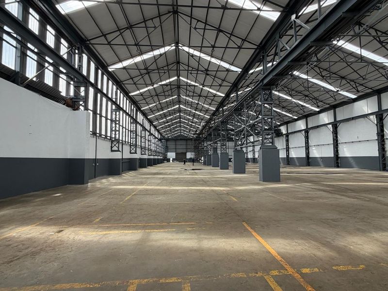 Expansive industrial warehouse space featuring high steel truss ceilings, abundant natural light from skylights, and durable concrete flooring, suitable for various commercial or industrial uses.