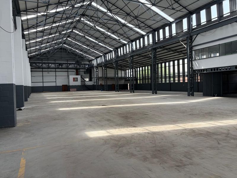Expansive industrial warehouse space featuring high ceilings with exposed steel trusses, concrete flooring, and abundant natural light from skylights and large windows.