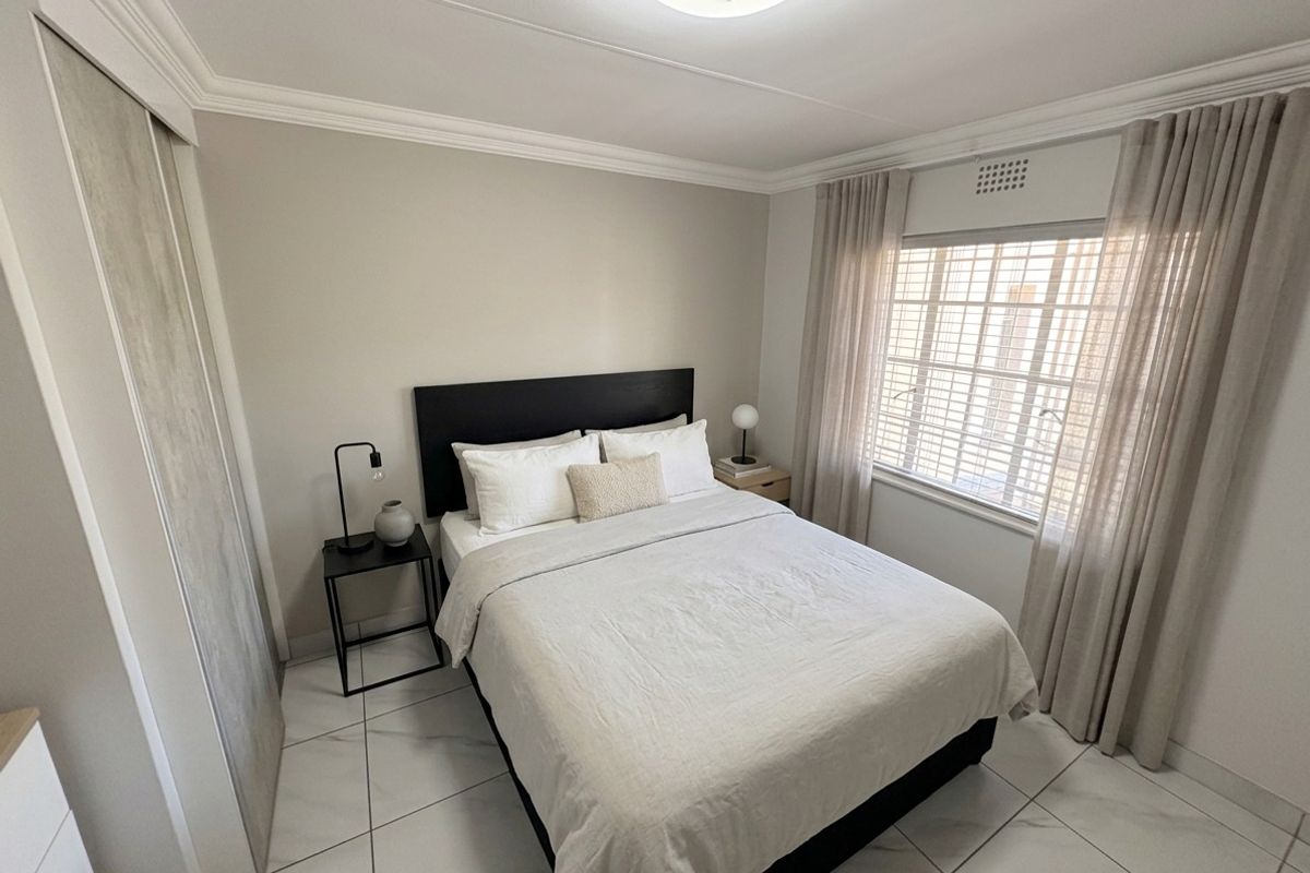 A well-maintained bedroom featuring neutral wall tones, tiled flooring, built-in wardrobe storage, and a window fitted with security bars.