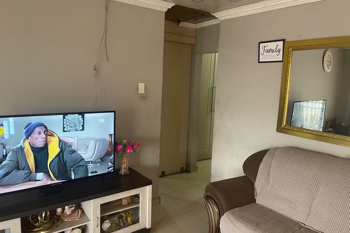 A living room area featuring tiled flooring and ceiling cornicing, currently showing signs of wear including damaged ceiling panels and scuffed wall finishes.