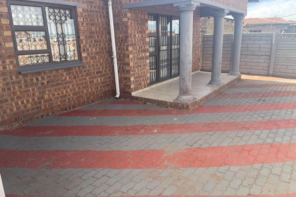 A rear exterior view featuring a face brick facade, a covered patio with concrete columns, and a patterned paved driveway area.