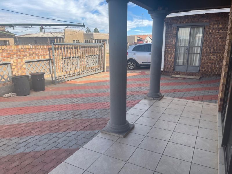 A covered patio area featuring tiled flooring and concrete columns, overlooking a paved driveway with a secure metal gate and brick boundary wall.