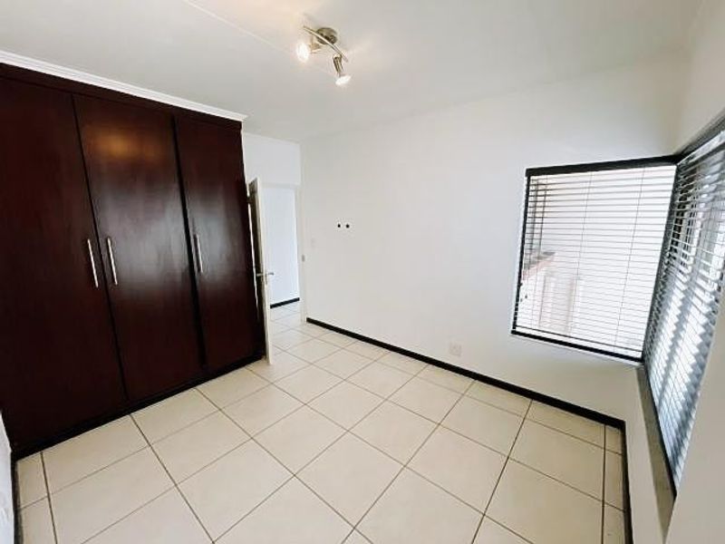 A bright, empty bedroom featuring tiled flooring, built-in dark wood cupboards, and venetian blinds on the windows.