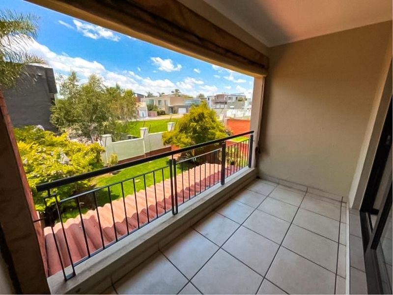 A covered balcony featuring tiled flooring and a metal safety railing, offering an open view of the surrounding neighborhood and garden area.