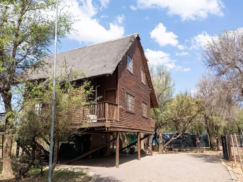 A rustic elevated log cabin featuring a traditional thatched roof, timber decking, and staircase, set within a natural farm landscape.