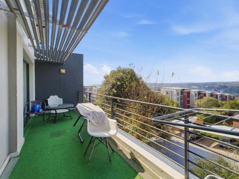 A spacious balcony featuring a metal balustrade, overhead slatted pergola, and artificial grass flooring, offering elevated views of the surrounding neighborhood.