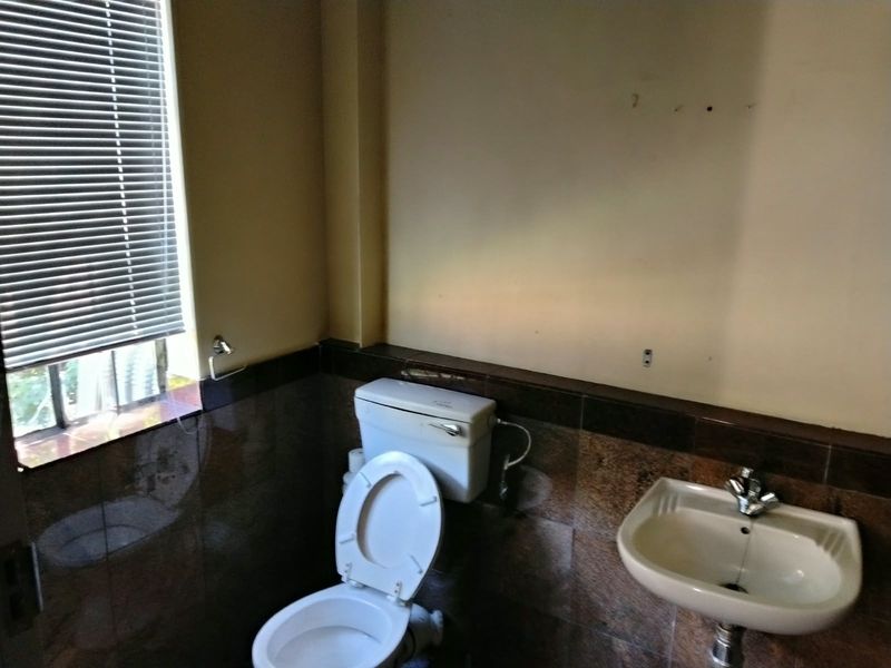 A compact industrial-style bathroom featuring a wall-mounted basin, standard toilet, and dark tiled wall finishes with venetian blinds.