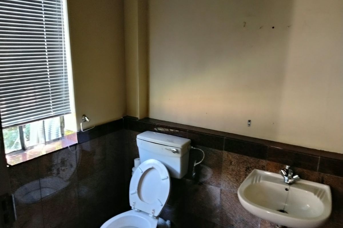 A compact industrial-style bathroom featuring a wall-mounted basin, standard toilet, and dark tiled wall finishes with venetian blinds.