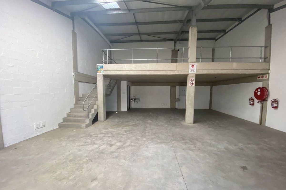 Spacious industrial warehouse unit featuring a concrete mezzanine level, staircase, exposed steel roof structure, and basic utility sink, suitable for storage or workshop use.