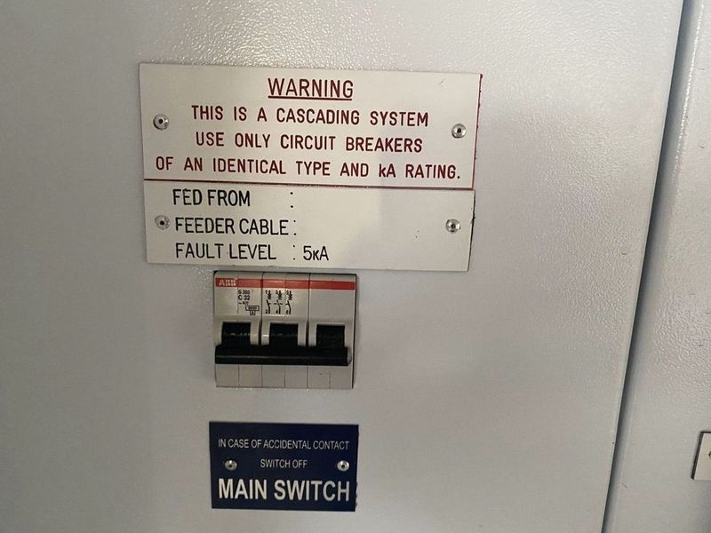 Industrial electrical distribution board featuring a main circuit breaker and safety warning signage for a cascading electrical system.
