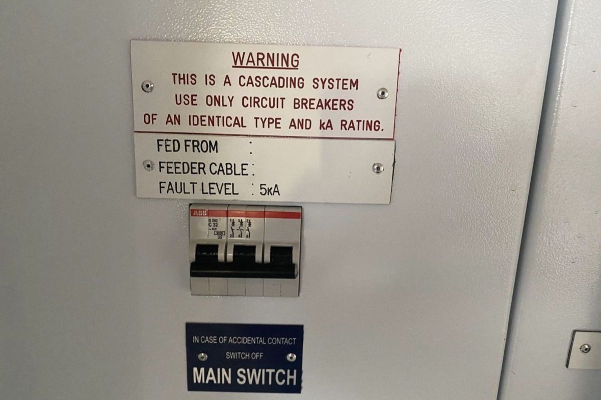 Industrial electrical distribution board featuring a main circuit breaker and safety warning signage for a cascading electrical system.