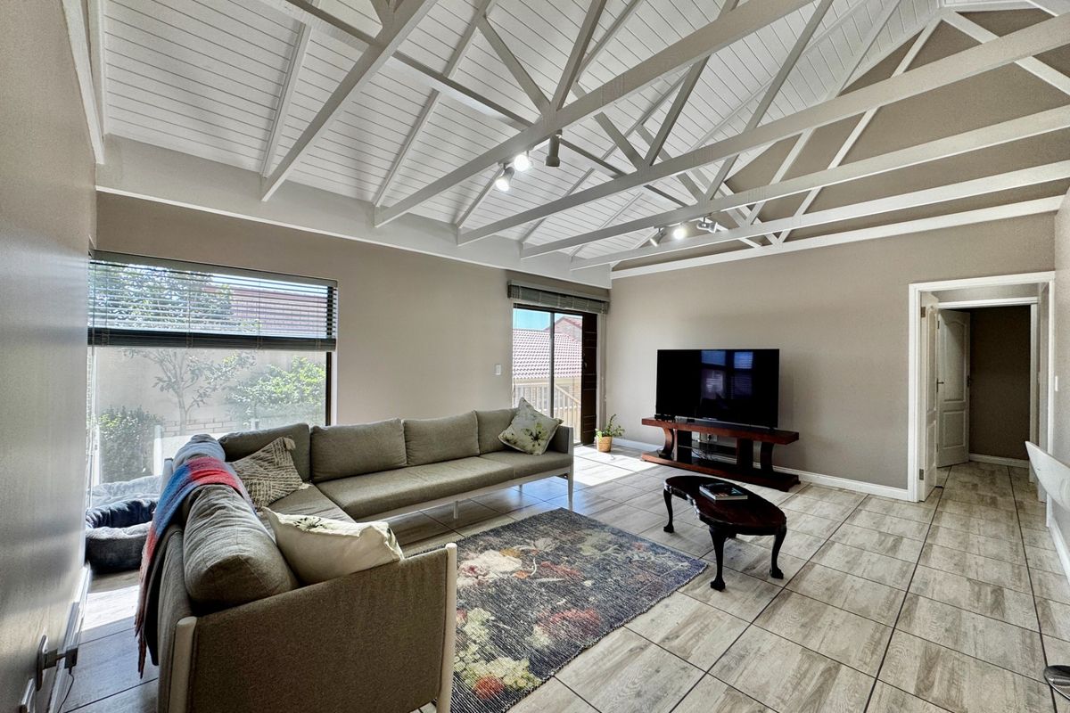 Spacious living room featuring high exposed timber truss ceilings, neutral wall tones, and large windows providing ample natural light with direct access to the exterior.