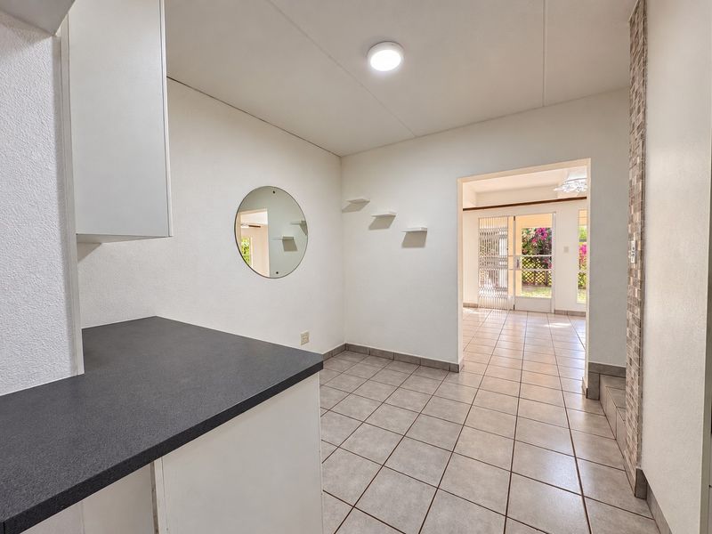 A clean, empty kitchen area featuring tiled flooring, built-in cabinetry with dark countertops, and a decorative tiled feature wall, leading into an adjacent living space.