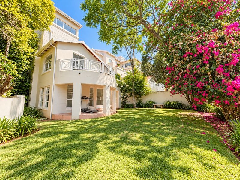 A well-maintained rear exterior view featuring a lush garden, a covered patio area, and an upper-level balcony with white railings.