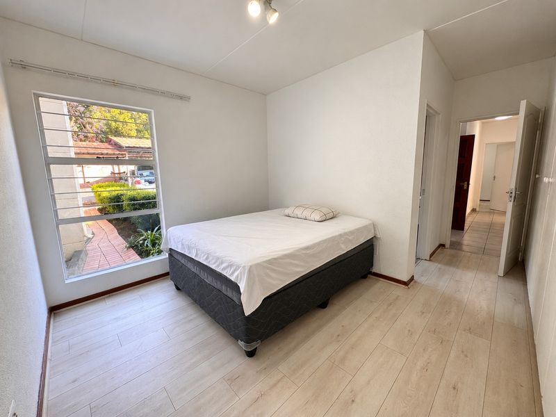 A bright bedroom featuring light-toned laminate flooring, a large window with security bars, and neutral wall finishes.