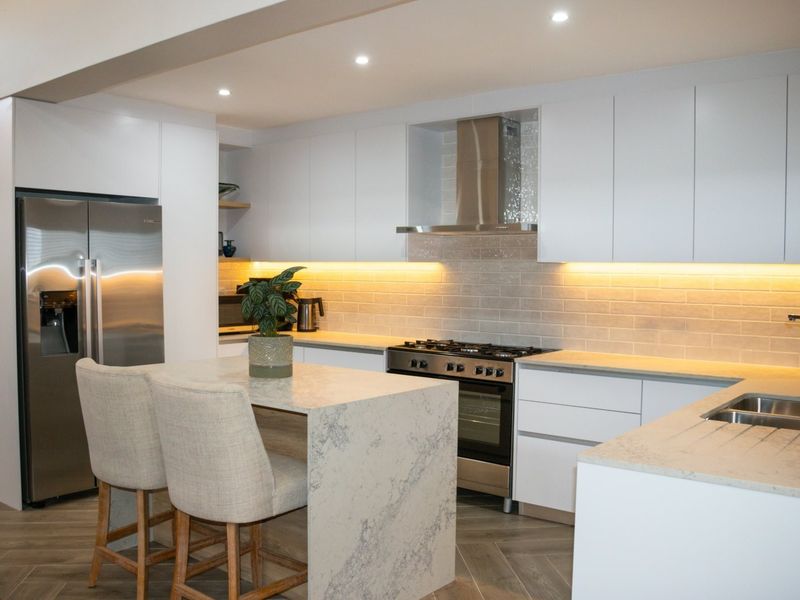 Modern kitchen featuring sleek white cabinetry, stone countertops, a gas stove with extractor fan, and elegant subway tile backsplash with integrated under-cabinet lighting.
