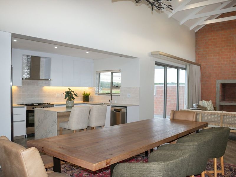Spacious open-plan living and dining area featuring a modern kitchen with stone island, vaulted ceilings with exposed trusses, and a feature brick fireplace.
