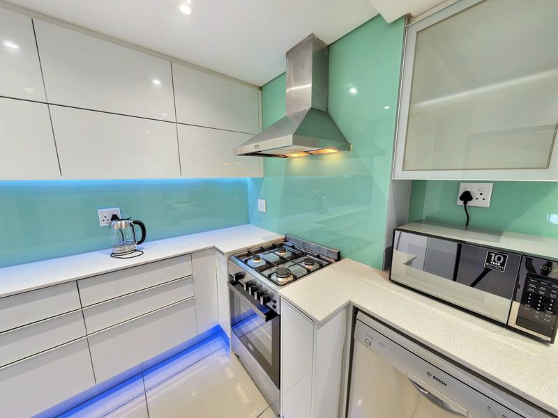 Modern kitchen featuring high-gloss white cabinetry, stone countertops, a vibrant glass splashback, and integrated stainless steel appliances with ambient under-cabinet lighting.