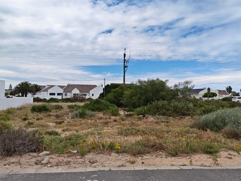 A vacant residential plot featuring natural coastal vegetation, situated in a developed neighborhood with established housing and utility infrastructure.
