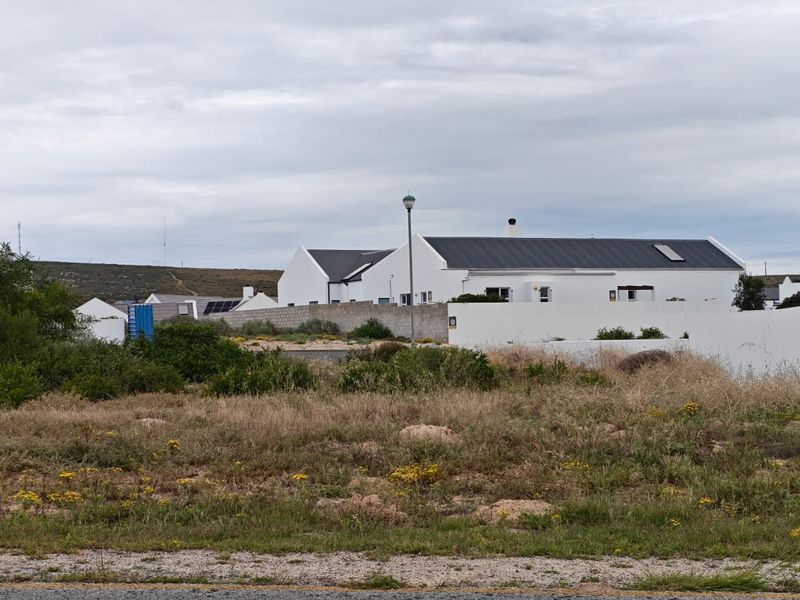 A view of a vacant residential plot with natural vegetation, featuring a boundary wall and surrounding residential development in the background.